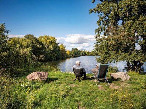 Zwei Menschen sitzen auf Stühlen am Ufer eines Flusses, umgeben von Bäumen und Wiese.