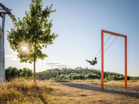 Person schaukelt an einer großen roten Schaukel auf einer Wiese, Baum links, Hügel im Hintergrund.