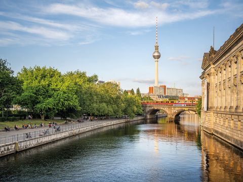 Spree mit Uferpromenade, Bäumen, Brücke und Fernsehturm im Hintergrund.