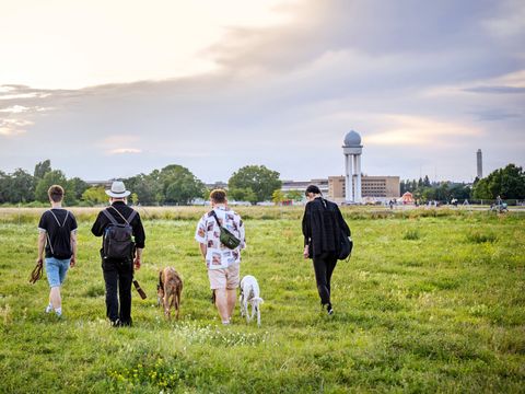 Vier Menschen mit zwei Hunden gehen über eine Wiese des Tempelhofer Felds, im Hintergrund der Radarturm und Bäume.