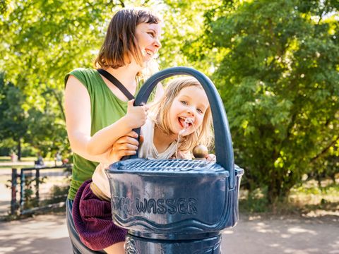 Frau und Kind lachend an einem blauen Wasserspender im Park.