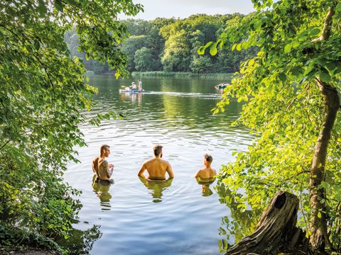 Drei Personen stehen im Wasser eines von Bäumen umgebenen Sees, im Hintergrund ein Boot.