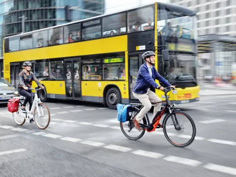 Zwei E-Bikes fahren auf einem Radweg. Dahinter fährt ein gelber Bus. Im linken Rand des Bildes sieht man die Kühlerhaube eines fahrenden Autos.