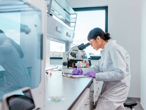 Eine weibliche Wissenschaftlerin schaut durchs Mikroskop und macht Notizen