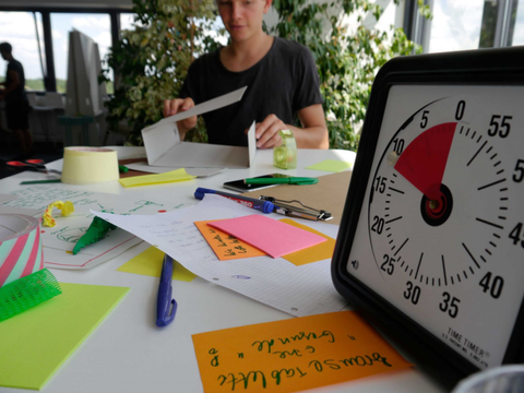 Tisch in einem Büroraum. Auf ihm befinden sich Büromaterialien und vordergründig eine Stopuhr. Die Gründungsteams erhalten Büroräume, Coaching und finanzielle Unterstützung.