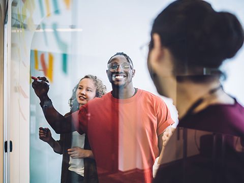 Eine Frau schaut in eine Spiegelung, die zwei Personen beim Beschriften eines Flipcharts zeigt