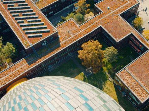 Luftaufnahme der Gebäude der Freien Universität Berlin mit begrünten Dächern. Im Vordergrund ist ein großes, kugelförmiges Dach mit einem Muster aus grünen und weißen Kacheln zu sehen. Die Gebäude sind von Bäumen und Grünflächen umgeben, was eine nachhaltige und grüne Campusumgebung betont.