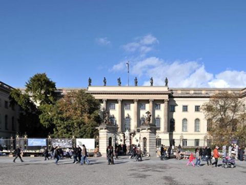 Das Bild zeigt das Hauptgebäude der Humboldt-Universität zu Berlin. Das prächtige Bauwerk besitzt eine beeindruckende Fassade mit Säulen und Statuen auf dem Dach. Vor dem Gebäude erstreckt sich ein großer Platz, auf dem zahlreiche Menschen spazieren gehen und sich aufhalten. Einige Bäume flankieren das Gebäude, und der Himmel ist klar mit einigen Wolken. Die Szene vermittelt einen lebhaften, geschäftigen Eindruck eines typischen Tages an der Universität.