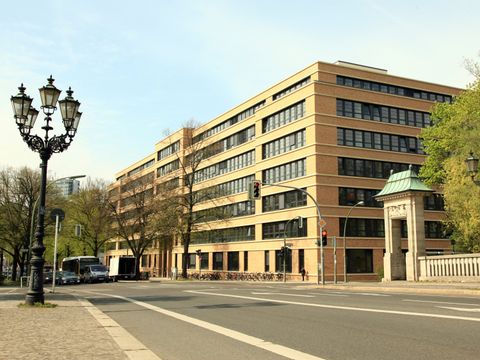 Ansicht eines modernen mehrstöckigen Gebäudes aus gelbem Backstein, das zur Technischen Universität Berlin gehört. Im Vordergrund ist eine Straße mit einem historischen Laternenpfahl und einem Torbogen zu sehen. Bäume umgeben das Gebäude, und der Himmel ist klar und blau.