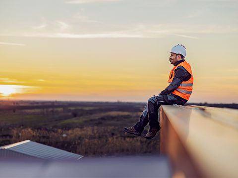 Person mit Warnweste und Bauhelm auf Dach schaut in die Ferne, Sonnenaufgang
