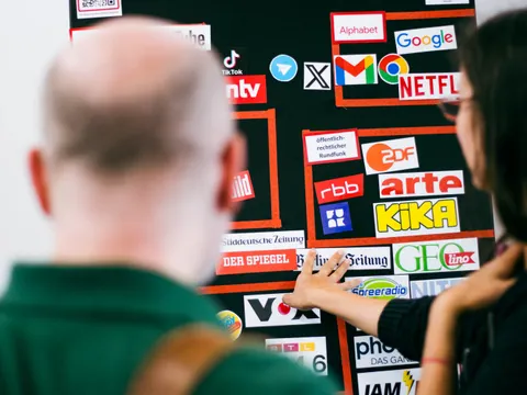 Zwei von hinten fotografierte Personen btrachten eine Wandbald mit verschiedenen Logos von Medienhäusern