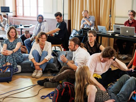 Menschen sitzen bei einer Veranstaltung auf dem Boden und hören einer Person zu, die ein Mikro hält.