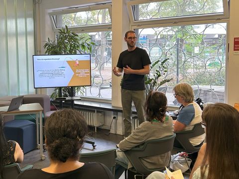Person steht neben Bildschirm und erklärt dem Publikum etwas