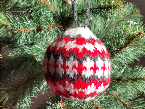 Fotografie einer gestrickten Weihnachtskugel, die an einem Nadelzweig hängt.