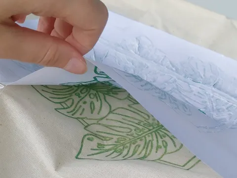 Eine Hand entfernt eine in Lavelöl getränkte Schablone, mit der eine florale Form auf einen Jutebeutel gedruckt wurde