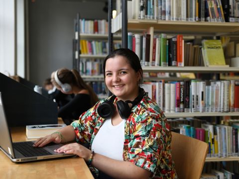 Eine Mädchen sitzt in einer Bibliothek an einem Laptop und lächelt in die Kamera