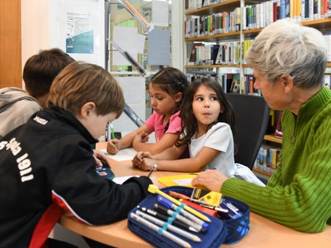 Kinder sitzen mit einer Erwachsenen am Tisch und zeichnen und schreiben