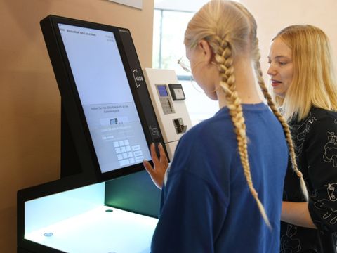 Ein Mädchen leiht zusammen mit einer Bibliothekarin ein Buch am Automaten aus