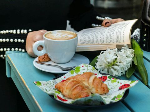 Eine Person sitzt an einem Tisch und liest ein Buch während auf dem Tisch ein Frühstück mit Getränk und Croissant steht