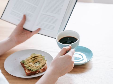 Eine Person liest ein Buch und hält in der anderen Hand einen Kaffee, auf dem Tisch steht ein Avocadobrot
