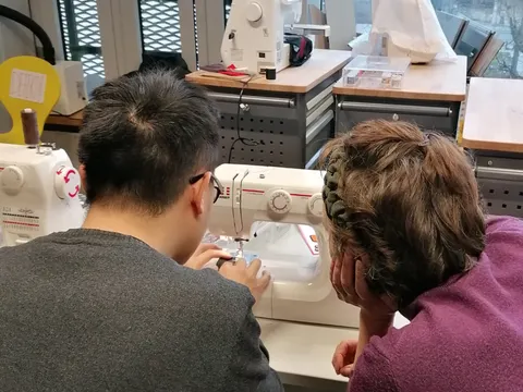 ein Mann sitzt an einer Nähmaschine und jemand guckt zu, beide sind von hinten zu sehen