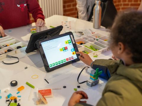 Kind sitzt an einem Tisch und bedient ein Tablet mit farbigen Symbolen, auf dem Tisch liegen Bauteile und Werkzeuge für ein Technikprojekt