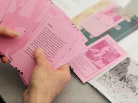 Hände halten einen Stapel rosa Post-it-Zettel mit Text, daneben liegen weitere rosa Zettel und schwarz-weiße Fotos auf einem Tisch