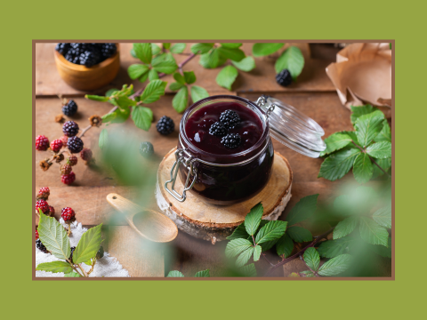 Ein Glas mit dunkler Brombeermarmelade auf einem Holzscheibenuntersetzer umgeben von Brombeerzweigen und einzelnen Brombeeren auf einem Holztisch