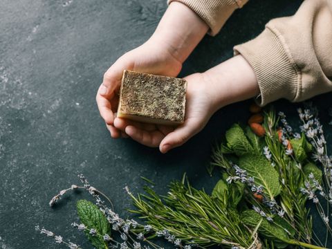 Hände halten ein rechteckiges Stück Seife mit getrockneten Blüten auf der Oberfläche neben frischen Wildkräutern auf dunklem Untergrund