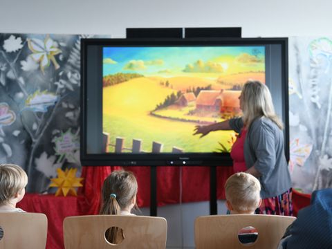 Frau präsentiert Kindern ein Bilderbuch auf einem großen Bildschirm.