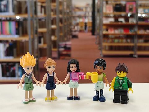Fünf kleine Spiezeug-Menschen stehen in der Bibliothek. Sie haben sehr unterschiedliche Haare, Kleidung und Hauttöne.
