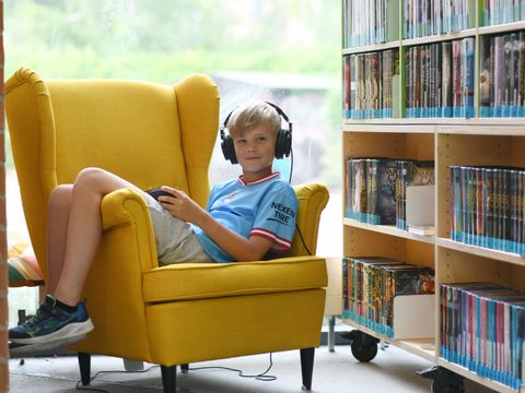 Ein Junge sitzt im Sessel in einer Bibliothek und hat Kopfhörer auf dem Kopf und ein iPad in der Hand