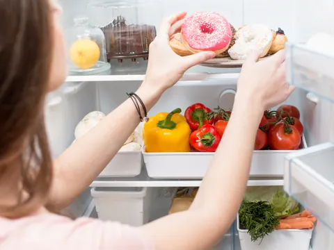 Ein offener Kühlscrhank mit Gemüse und Gebäck. Eine Frau nimmt sich Donuts.