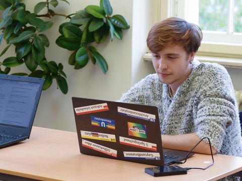 Junge Erwachsene mit einem Laptop in der Bibliothek. Auf dem Laptop sind viele Queer-Pride-Aufkleber.