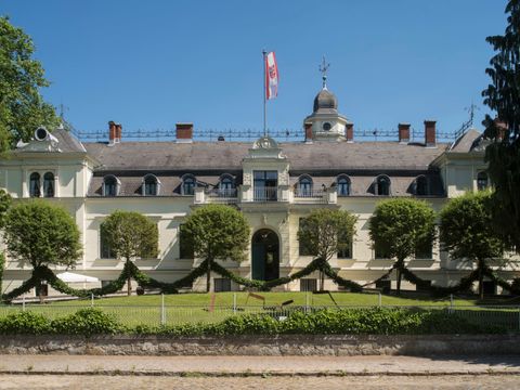 Außenansicht des Schloss Britz. Ein weißes Gebäude. Auf dem Dach weht die Berlin Flagge. Vor dem Schloss eine Wiese.