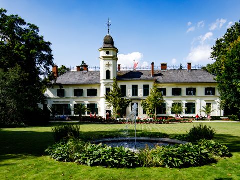 Das Schloss Britz auf einer grünen Rasenfläche. Ein weißes Haus mit vielen Fenster mit grünen Fensterläden. In der Mitte des Hauses ein schmaler Turm mit einem spitzen Dach
