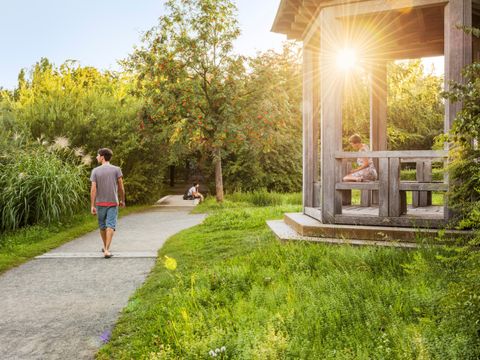 Ansicht eines Parks. Auf einer Wiese rechts steht ein Holzpavillion. Eine Frau sitz darin und ließt ein Buch. Ein Mann geht auf dem Weg spazieren. Die Sonne scheint