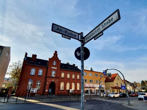 Im Vordergrund eine Straßenkreuzung. Auf der Ecke steht ein Straßenschild. Nach links zeigt das Schild "Alt-Rudow" nach rechts das Schild "Köpenicker Straße". Im Hintergrund ein rotes Backsteinhaus- die alte Dorfschule Rudow. Daneben ein Haus mit Flachdach in orange. Der Himmel ist blau mit leichten Schleierwolken.