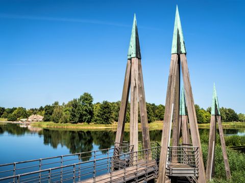 Im Bildvordergrund sind mehrere hölzerne Pfähle zu sehen. Immer drei Pfähle laufen oben zusammen und bilden eine große Stütze. Die drei Pfähle sind oben mit grüne Kappen verbunden. Zwischen den Pfählen verläuft ein Weg, der aus einzelne Holzplanken besteht. Es ist eine Brücke. Die Brücke führt über einen See, den man im Hintergrund erkennt. Auf der anderen Seite des Sees erkennt man schon das Ufer. Es ist bewaldet. Auf der linken Seite des Ufers sieht man ein graues Gebäude