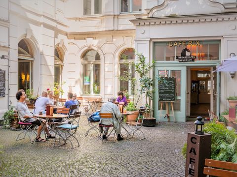 Eingangsbereich des Heimtahafens Neukölln von Außen. Kopfsteingepflasteter Innenhof. Auf Stülen an Tischen sitzen im linken Bildteil Personen und trinken Kaffee. Über eine mintfarbene Eingangstür gelangt man ins Innere des Heimathafens. Oberhalb der Tür befinden sich die Logos des Heimthafens Neukölln. Ein Bug eines Schiffes und darunter der weiße Schriftzug Heimathafen Neukölln auf schwarzem Hintergrund. Oberhalb der Schriftzug Café Rix in einzelnen goldenen Buchstaben. Die Hausfassade des Heimathafens ist hell. Die Sonne scheint.