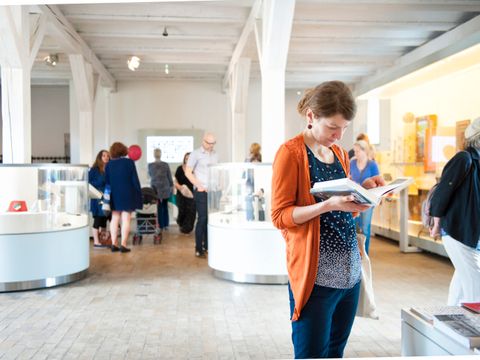 Auf der rechten Bildseite eine Frau, die gerade ein Buch in den Händen hält und darin ließt. Die Frau hat eine blaue Jeans an und ein blaues Shirt, mit weißen Applikationen am Ende des Shirts. Sie trägt eine orangefarbene Strickjacke darüber. Sie hat braune Haare, die am Hinterkopf zusammen gebunden sind. Im Hintergrund ein weißer Raum mit mehreren weißen, halbhohen Vitrinen. Hierin sind verschiedene Objekte untergebracht