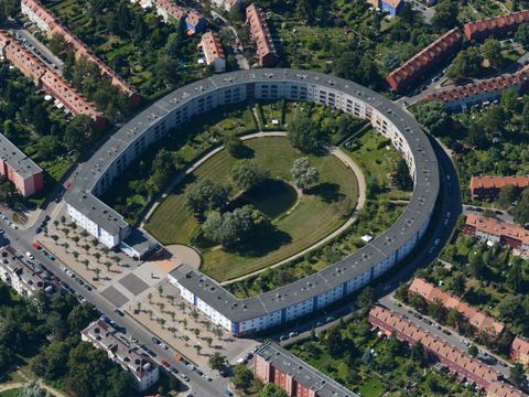 Luftbildaufnahme der Hufeisensiedlung. In der Mitte des Bildes die Hufeisensiedlung mit einem grauen Dach. Der Innenhof der Siedlung ist begrünt und es gibt einen kleinen Teich in der Mitte. Rechts und links, sowie oberhalb der Hufeisensiedlung gehen strahlenförmig weitere Reihen Häuser weg, von denen nur die roten Dächer zu sehen sind