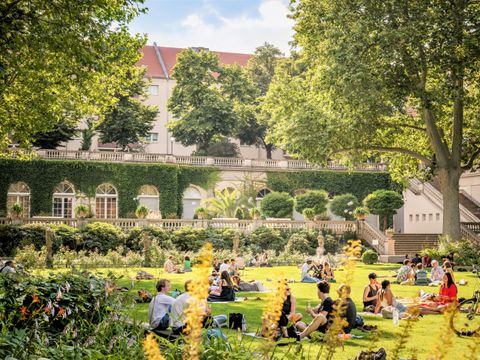 Menschen sitzen in kleinen Gruppen auf einer Wiese. Rechts sieht man einen Baum. Im Hintergrund ein Laubengang. Die Sonne scheint