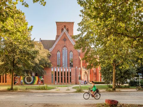 In der Mitte des Bildes eine rote Backsteinkirche. Rechts und links davon Bäume. Davon fährt jemand auf einem Fahrrad vorbei.