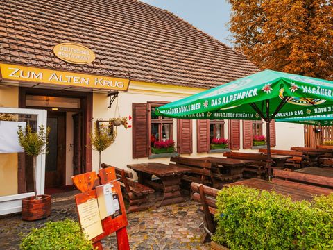 Auf der rechten Bildseite eine niderige grüne Häcke und daneben ein grüner Sonnenschirm. Dahinter viele Holzbänke und Tische aus dunklem Holz. Dahinter ein niedriges Haus mit einer hellgelben Fassade und einem rot-braunen Dach. Über dem Eingang ein Schild mit der Schrift "zum Alten Krug". Weiße Buchstaben auf senfgelben Untergrund
