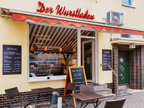 Gelbe Hausfassade mit einem großen Schaufenster und daneben einer geöffneten glässernen Eingangstür. Drei Stufen führen zu dem Ladengeschäft hoch. Links von den treppen steht eine Aufstelltafel. Davor zwei Tische mit jeweils zwei Stühlen. Über dem Schaufenster ein roter verschlungener Schriftzug "Der Wurtsladen"