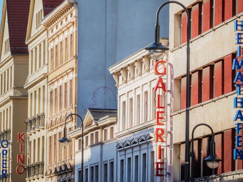 Helle Häuserfassaden mit den seitlich am Haus angebrachten, senkrecht verlaufenden Schriftzügen "Heimathafen", "Galerie", "Kino" und "Oper". Die Schriftzüge sind abwechselnd blau und rot und bestehen aus einzelnen Buchstaben