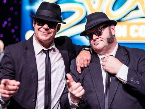 Zwei Männer in dunklen Anzüge stehen Schulter an Schulter. Sie haben beide einen schwarzen Hut auf. Im Hintergrund lässt sich der Schriftzug "Stars in Concert" erkennen. Gelb-orangene Schrift auf blauem Hintergrund.
