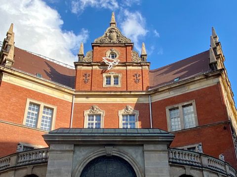 Das Bild zeigt ein Backsteingebäude. Im unteren Teil des Gebäudes der Eingang. Dieser Teil ist heller Sandstein. Das restliche Haus besteht aus rotem Backstein. Das dach hat leicht dunkelrote Dachschindel. Der Eingangsbereich ist eine Durchfahrt. Darin eine schmiedeeiserne Tür. Darauf der Schriftzug "Alte Post" in weißen einzelnen großen Buchstaben. Hinter dem Sandsteinteil beginnt das restliche Haus. Es hat am Dach mehrere kleine gemauerte Spitzen und im Dachgiebel eine stilisierte Taube, die leuchten kann. Der Himmel ist blau. Auf der linken Seite sieht man eine weiße Wolke