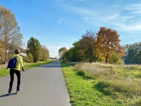Ein asphaltierter Weg in der Bildmitte. Eine Frau ist auf diesem Weg mit Inlinern unterwegs. Man sihet sie von hinten. Sie hat eine gelbe Jacke an und eine schwarze Hose. Sie hat einen schwarzen Turnbeutel auf dem Rücken und hat kurze blonde Haare. Rechts und links des Weges ist Wiese. Auf der Wiese wachsen Bäume, deren Blätter schon in rot, gelb und braun herbstlich eingefärbt sind. Der Himmel ist blau mit leichten Schleierwolken